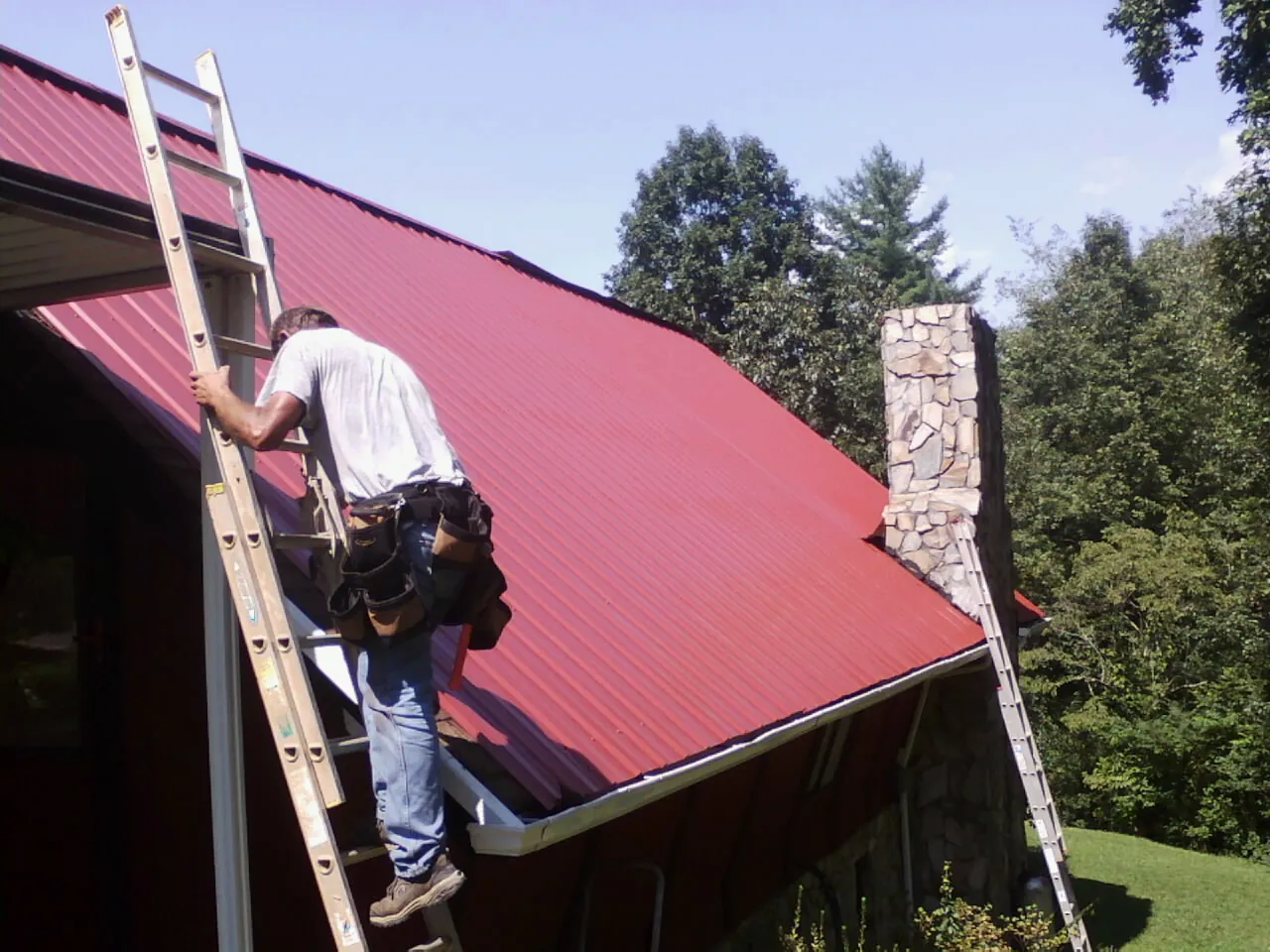 Roofer on a ladder beside a red metal roof and stone chimney — Soffit & Fascia Repair by Roofing Brothers in Fort Ogden